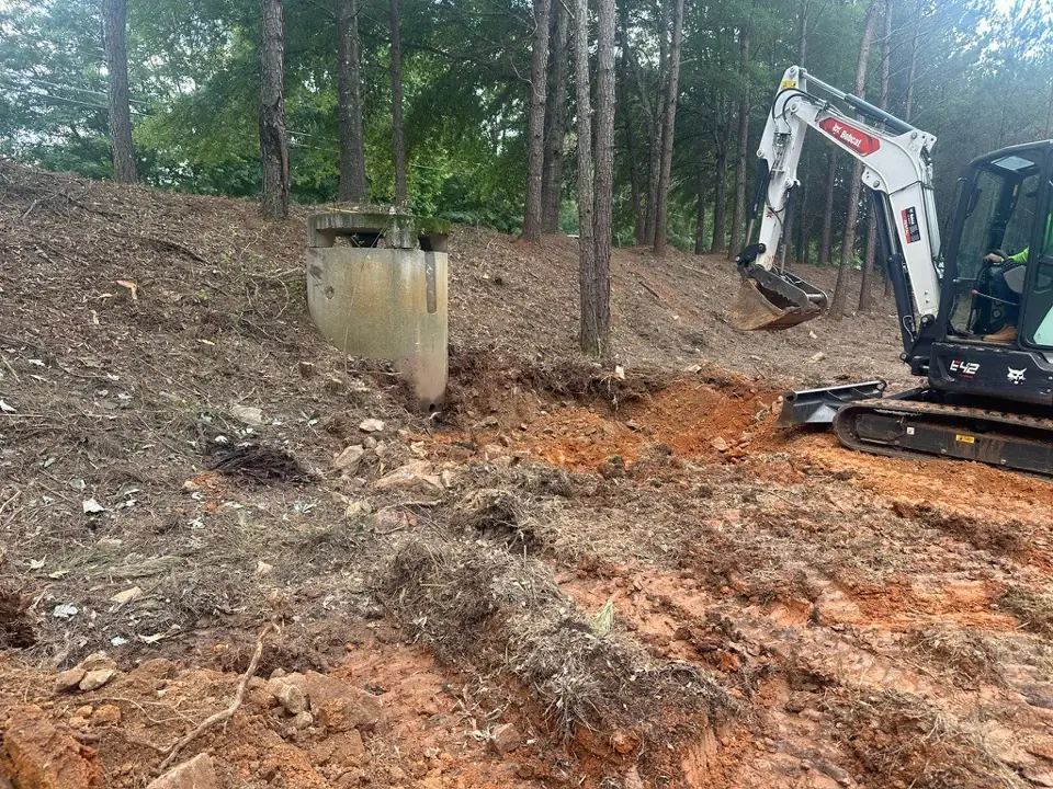 A mini excavator digging out eroded soil around a concrete stormwater outlet structure on a wooded slope